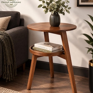 Peut inclure: Une table d'appoint ronde en bois avec trois pieds inclinés et une étagère circulaire inférieure. La table supporte un vase sombre cannelé avec du feuillage vert et deux livres empilés intitulés The Kinfolk Home et Live Beautifully.