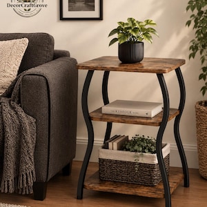 May include: A three-tier side table with a dark wood grain finish and curved black metal legs. The top shelf holds a potted plant in a ribbed black container. The middle shelf holds a book titled The Kinfolk Home. The bottom shelf holds a woven basket.