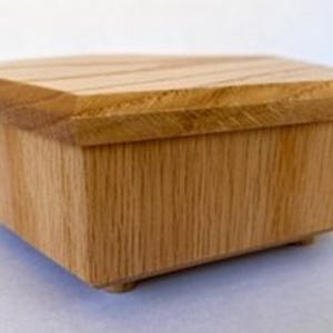 May include: A rectangular wooden box with a natural light brown grain finish. The box features a flat, slightly overhanging lid with beveled edges and rests on four small, low-profile feet at the base, showcasing a simple, clean construction.