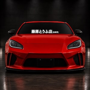 Puede incluir: Un coche deportivo de color rojo brillante visto desde el frente, con una calcomanía blanca en el parabrisas que muestra el texto japonés "藤原とうふ店(自家用)". El vehículo tiene faros angulares, una rejilla frontal negra y espejos laterales.