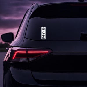 May include: A vertical rectangular white sticker applied to the rear window of a black vehicle. The sticker features black Japanese kanji characters arranged in a single column, reading 悲しい少年, which translates to sad boy.