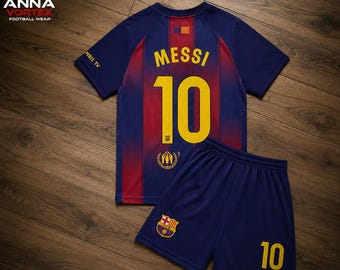 Puede incluir: Un conjunto de fútbol azul y rojo que incluye camiseta y pantalón corto, con el nombre MESSI y el número 10 en amarillo. La camiseta tiene rayas verticales, un pequeño detalle de bandera en el cuello y un logotipo en el pantalón. El texto AMBIL TV está en la manga.