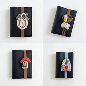 Pode incluir: Quatro cadernos pretos com elásticos. O primeiro apresenta um aplique de relógio com renda. O segundo tem um buquê de flores de tecido. O terceiro mostra uma casa em forma de cogumelo. O quarto mostra uma bolsa xadrez azul com o texto Apple day.