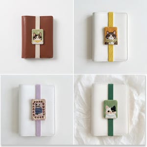 May include: Four rectangular notebooks, each secured with a vertical elastic band featuring a rectangular embroidered patch of a cat. The notebooks are brown or white, with elastic bands in cream, yellow, lavender, or green, and cat designs.