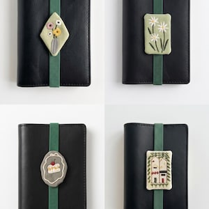 Puede incluir: Cuatro cuadernos negros, cada uno asegurado con una banda elástica verde que presenta un parche bordado único. Los parches muestran un jarrón con flores, margaritas blancas, un trozo de pastel con una cereza y una casa pequeña.