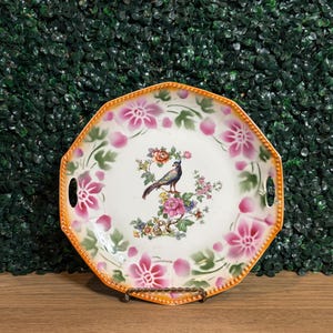 May include: An octagonal porcelain serving dish with an orange beaded rim and two integrated side handles. The center features a colorful bird perched on a floral branch, surrounded by a border of large pink flowers and green leaves on white.