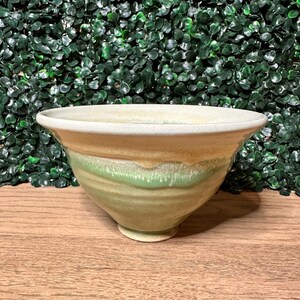 May include: A ceramic bowl with a wide, flared rim and a tapered base. The exterior features a layered glaze with a creamy off-white top section that transitions into a soft, muted green hue, showing visible drip patterns along the center body.