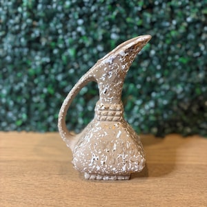 May include: A tan ceramic pitcher with a textured, speckled white finish. The vessel features a square base, a narrow neck with a decorative grid-patterned band, and a single curved handle extending from the side to the top of the spout.