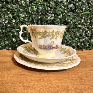 May include: A porcelain teacup and saucer set featuring a cream-colored base with a delicate green vine and leaf border. The cup displays a small illustration of a mouse wearing a blue jacket, while the saucer mirrors the vine pattern.