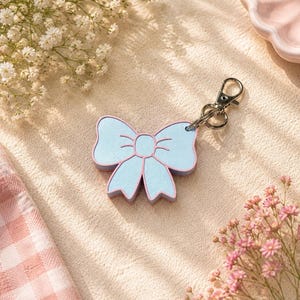 May include: A light blue bow-shaped keychain with a pink outline and a silver-toned metal lobster clasp. The bow features a textured surface and a central knot detail, attached to a circular split ring for securing keys or other small items.