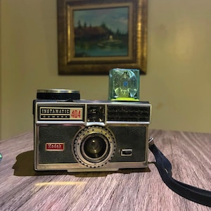 Peut inclure: Un appareil photo vintage Kodak Instamatic 404 avec un corps texturé gris foncé et des accents métalliques argentés. L'appareil présente un objectif central, un logo Kodak rouge et un cube flash bleu translucide monté sur le dessus.