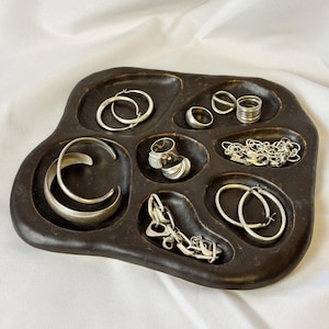 May include: A dark brown, organic-shaped jewelry tray with multiple recessed compartments containing various silver-toned items, including hoop earrings, a cuff bracelet, several rings, a chain necklace, and a link bracelet.