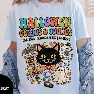 May include: A light blue t-shirt featuring a graphic print with the text HALLOWEN CURIOS & ODDIES EST. 1978 | HANDCRAFTED | BO-TIQUE. The design includes a black cat with a bow tie, a skeleton, a ghost, a bat, and signs reading VINTAGE FINDS & SPOOKY GOODS and ALL GOOD THINGS.