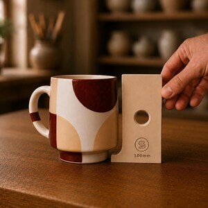 May include: A ceramic mug featuring a geometric pattern of dark red, light tan, and cream shapes, positioned next to a beige rectangular measuring tool with a circular cutout and the text S 100mm, indicating a height of approximately 3.94 inches.