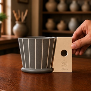 May include: A dark gray ceramic coffee dripper with vertical white stripes and a light beige handle attachment. The handle features a circular cutout, a circular logo with the letter S, and the text 100mm printed on the lower section.