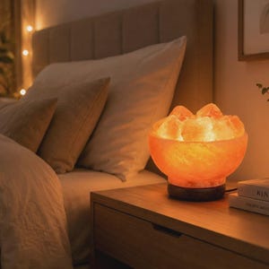 May include: A bowl-shaped Himalayan salt lamp with a warm orange glow, featuring rough-hewn salt chunks inside a rounded vessel. The lamp sits on a dark circular wooden base, emitting a soft, ambient light that illuminates the surrounding area.