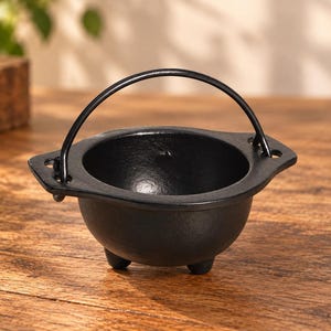 Pode incluir: Um pequeno caldeirão de ferro fundido preto com uma taça arredondada, três pernas curtas e uma alça de metal fina fixada a duas abas laterais. O interior é liso e escuro, enquanto o exterior apresenta um acabamento texturizado de ferro.