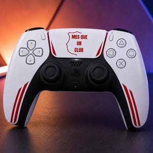 Peut inclure: Une manette de jeu vidéo sans fil blanche et noire avec des rayures rouges sur les poignées et le texte "MES QUE UN CLUB" imprimé en rouge sur le pavé tactile central, accompagnée des boutons directionnels et des deux joysticks analogiques.