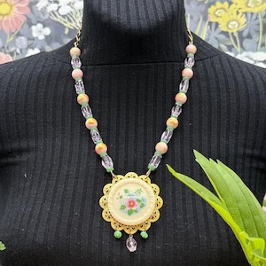 May include: A necklace featuring a round, cream-colored pendant with a floral design, framed by a gold-tone scalloped metal border. The chain consists of alternating clear faceted beads, round pink beads, and small green spacers, with a single clear bead drop.