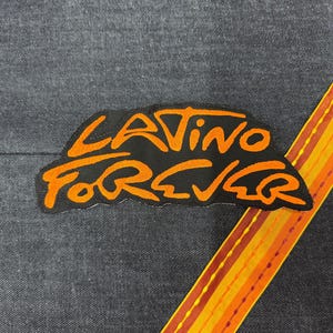 May include: A black fabric patch featuring the orange embroidered text "LATINO FOREVER" in a stylized script font. The patch is positioned on a dark gray denim fabric next to a diagonal strip of orange and yellow fabric with visible stitching.