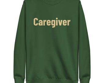 Puede incluir: Una sudadera de manga larga color verde bosque con la palabra Caregiver impresa en una fuente sans-serif de color beige claro en el centro del pecho. La prenda tiene cuello redondo, puños acanalados y un dobladillo acanalado del mismo color.