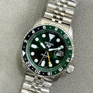May include: A Seiko 5 Sports automatic GMT watch with a dark green dial, luminous hour markers, and a silver-tone stainless steel jubilee bracelet. The watch features a two-tone black and green rotating bezel, a date window, and a yellow GMT hand.