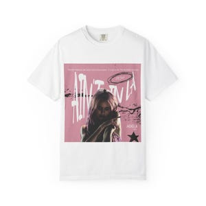 Può includere: Una t-shirt bianca a maniche corte con una stampa grafica rosa centrale. Il design include il ritratto di una persona con i capelli rosa, il testo "AIN'T IN LA", "PRIMA" e "ADÉLA", insieme a un'icona a stella nera e texture nere usurate.