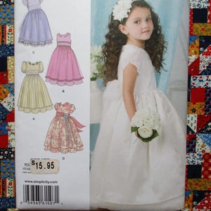 Puede incluir: Una niña con un vestido blanco sostiene un ramo de flores blancas, junto a un patrón de vestidos infantiles. El patrón muestra ilustraciones de varios diseños de vestidos en colores pastel. Una colorida colcha de retazos está en el fondo.