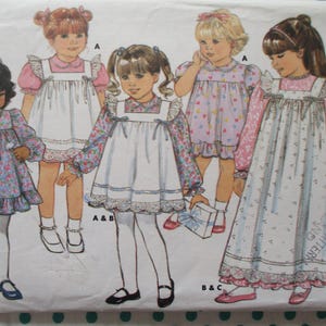 Op de afbeelding: Een vintage naaipatroon met illustraties van jonge meisjes in jurken. De jurken hebben bloemenprints, kanten randen en schortstijlen. Het patroon is gelabeld "CLASSICS" en bevat maatopties.