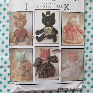 Könnte beinhalten: Ein Vintage-Schnittmuster mit dem Titel "Jiffy Six Pack" mit sechs verschiedenen Katzendesigns. Die Puppen sind in verschiedenen Outfits gekleidet, darunter Kleider, Hüte und Fliegen. Der Hintergrund ist ein weißer Stoff mit türkisfarbenen Punkten.
