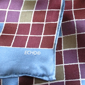 Puede incluir: Un pañuelo de seda con un diseño geométrico de cuadrados en tonos burdeos, morados y verde oliva, delineados en blanco. El borde es azul claro con el nombre de la marca "ECHO" impreso en blanco.