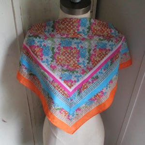 May include: A square scarf with a floral pattern in orange, pink, and blue. The scarf has a bright orange border, a pink inner border, and a light blue stripe. The scarf is displayed on a mannequin.