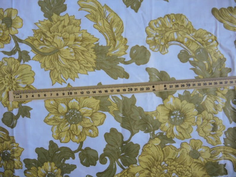 May include: White fabric with a repeating pattern of yellow flowers and green leaves. The fabric is measured with a ruler, showing the scale of the pattern.