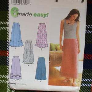 May include: A sewing pattern package featuring six skirt designs, with a woman modeling a pink skirt. The package has the text "6 made easy!" and includes illustrations of different skirt styles. The background is a plaid fabric.