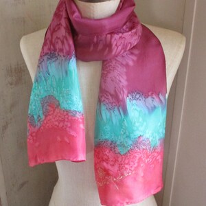 May include: A vibrant, hand-painted silk scarf in shades of fuchsia, turquoise, and coral. The scarf is draped over a mannequin, showcasing its flowing design. The bottom right corner features a signature in white script.
