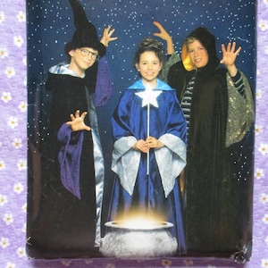 May include: Image of three children dressed in wizard costumes. One wears a black robe and hat, another a blue robe with a star wand, and the third a black hooded cloak. A cauldron with glowing light is in front.