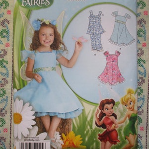 May include: A Simplicity sewing pattern featuring a young girl in a light blue fairy dress with wings. The pattern includes illustrations of fairy-themed dresses and outfits. The background has a floral pattern.