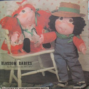 Puede incluir: Patrón de costura vintage con dos muñecos de tela esculpidos. Uno está sentado, con un vestido rojo y un gorro. El otro está de pie, con un mono y un sombrero de paja. El patrón se titula "Blossom Babies" de Fave Wine for McCall's.