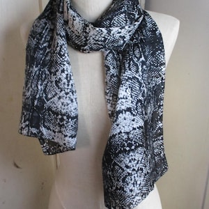 Puede incluir: Bufanda con estampado de piel de serpiente en blanco y negro, colocada sobre un maniquí. El patrón se repite en toda la tela. La tela parece ligera y fluida, un accesorio de moda.