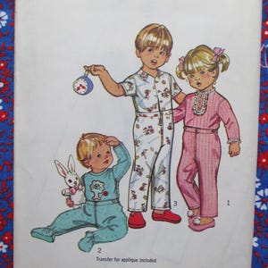 Può includere: Illustrazione di tre bambini in pigiama. Un bambino tiene in mano un orologio, un altro un coniglietto di peluche. I pigiami sono in vari colori e fantasie, tra cui un completo azzurro, un completo rosa e un completo bianco con un disegno floreale. Il testo "Transfer per applicazione incluso" è in basso.