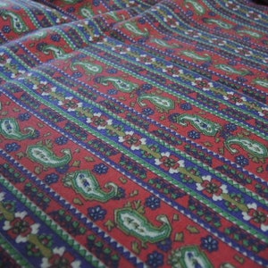 Op de afbeelding: Rode stof met een blauw, groen en wit paisley-patroon. De stof heeft een gestreept ontwerp met een herhalend patroon.