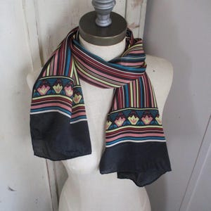 May include: A colorful scarf with vertical stripes in red, teal, yellow, and pink. The scarf has a black border and a repeating floral pattern in pink, yellow, and teal. The scarf is draped over a mannequin.