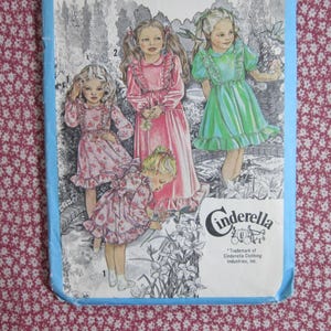 May include: A sewing pattern envelope titled Cinderella featuring illustrations of three children wearing dresses with ruffles and floral patterns. The artwork shows one child in a green dress and two in pink floral dresses, set against a garden background.