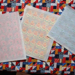 Peut inclure: Trois feuilles de papier à motifs. L'une présente un motif floral rose, une autre des fleurs orange et vertes répétées, et la troisième une bordure florale bleue. Les feuilles sont sur un tissu patchwork coloré.