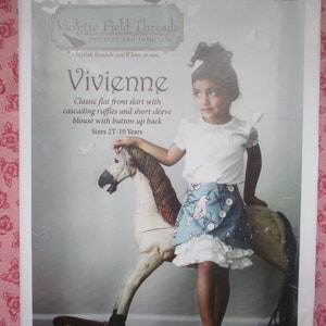 以下が含まれることがあります： 「Vivienne」という女の子のスカートとブラウスの縫製パターンです。パターンはViolette Field Threads製で、2T〜10歳のサイズに対応する説明書が含まれています。画像には、完成した衣装を着てロッキングホースに乗っている若い女の子が写っています。