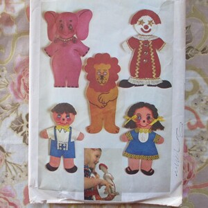 May include: A vintage sewing pattern featuring five felt characters: a pink elephant, a red clown, a brown lion, a boy with a blue shirt and overalls, and a girl with a blue dress. The pattern includes instructions for making the characters.