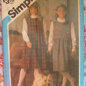 May include: A vintage Simplicity sewing pattern featuring two girls wearing dresses. The girl on the left is wearing a plaid dress with a white collar and the girl on the right is wearing a solid blue dress with a white collar. The pattern is for sizes 6-14.