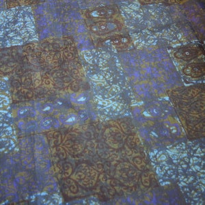 May include: A fabric with a patchwork design featuring various shades of blue, brown, and purple. The fabric has a repeating pattern of intricate floral designs.