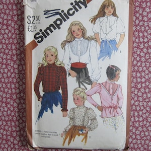 May include: A Simplicity sewing pattern envelope featuring illustrations of four different blouse designs with long or short sleeves, ruffles, and button-front closures. The cover displays the brand name Simplicity and price markings of $2.50 and £1.35.