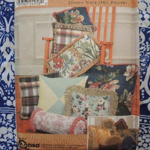 Könnte beinhalten: Eine Nahaufnahme eines Buchumschlags mit dem Text "Design Your Own Pillows". Der Umschlag zeigt eine Vielzahl von bunten Kissen mit Blumen-, geometrischen und karierten Mustern. Die Kissen sind auf einem Stuhl mit einem Holzrahmen angeordnet.